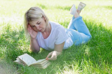 Genç sarışın bir kadın güneşli bir yaz gününde parkta çimlerin üzerinde uzanıyor ve bir kitap okuyor. Doğada eğlence ve mesafe öğrenimi.