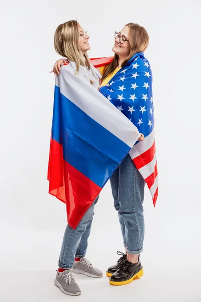 Young girls in American and Russian flags laugh and hug. International relations and friendship between states. White background. Vertical.