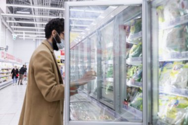 Büyük bir süpermarketin donmuş gıda reyonunda maskeli bir adam. Bej ceketli, sakallı, genç bir esmer. Koronavirüs salgını. Yan görüş. Bulanık.