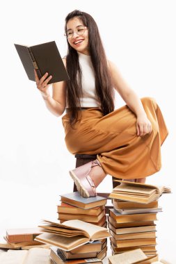 Gözlüklü genç kadın kitap okuyor. Uzun saçlı, gülümseyen esmer bir sürü kitapta oturuyor. Beyaz arka plan. Dikey.
