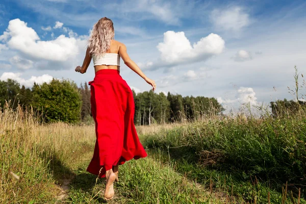 Young woman runs in the field. Blonde in long hair in a red skirt. Enjoy freedom in nature on a sunny summer day. Full height. Back view.