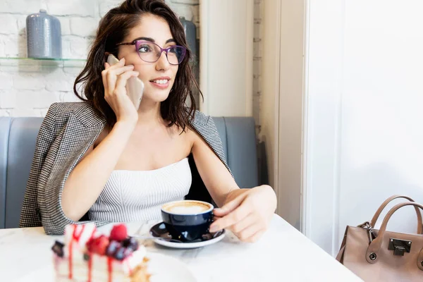 Smiling young woman with glasses sits in a cafe with a laptop, talking on the phone and looking out the window. Remote work and training.