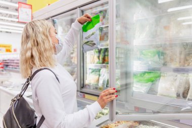 Donmuş gıda bölümünde tıbbi maske takan genç sarışın kadın. Bir salgın sırasında sağlık ve düzgün beslenme.