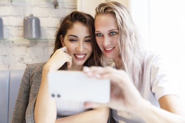 Kız arkadaşlar bir kafede masada otururken selfie çekerler. Gülümseyen genç kadınlar, sarışın ve esmer. Arkadaşlık ve ilişkiler.