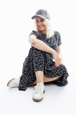 Elbiseli genç bir kadın oturuyor. Siyah elbiseli ve şapkalı gülümseyen şık bir sarışın. Beyaz arka plan. Dikey.