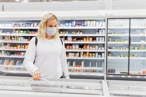 Молодая Женщина Маске Супермаркете Выбирает Замороженные Продукты Блондинка Рюкзаком Пандемия — стоковое фото
