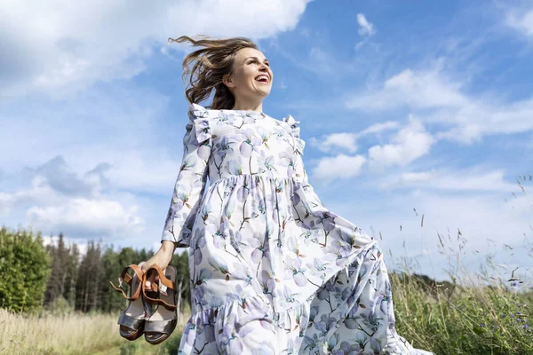 Güneşli bir günde, uzun romantik bir elbise giymiş, gülen genç bir kadın tarlada koşuyor. Mutluluk doğayla birliktir..