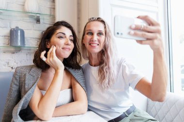 Kız arkadaşlar bir kafede bir masada oturup fotoğraf çekerler. Gülümseyen sarışın ve esmer.