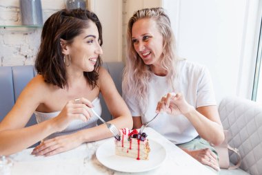 Kız arkadaşlar bir kafede bir masaya oturur ve pasta yerler. Gülümseyen sarışın ve esmer.