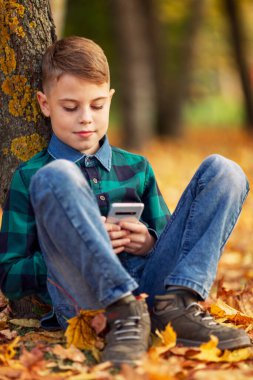 Kot pantolonlu bir çocuk bir sonbahar ormanında bir ağacın altında telefonuyla oturuyor. Dikey.