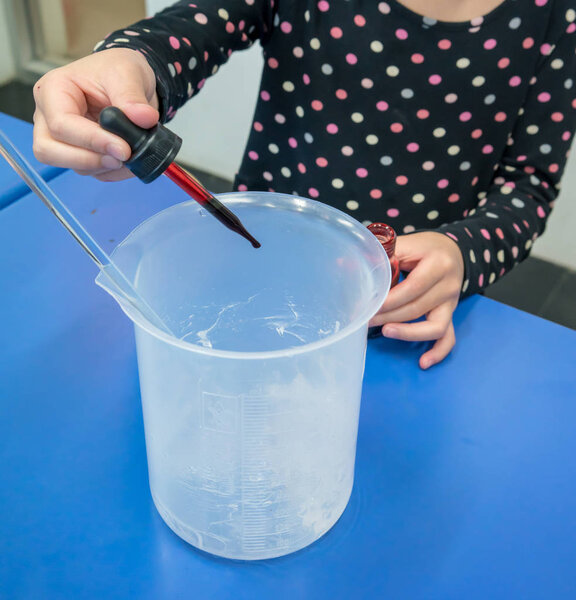 Girl using droplet to add red color into a plastic beaker on blue table. DIY Hand gel experiments for young scientist education.