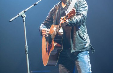 Sahnede akustik gitar çalan deri ceket giyen adam. 