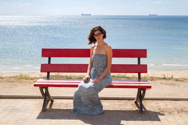 Kırmızı bir bankta oturan genç beyaz kadın modeli. Arka plan plaj ve deniz. Yatay olarak çerçeveli atış.