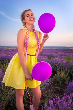 Uzun örgüler ile güzel bir model bir lavanta alanında duruyor. İki balon elinde tutar. Dilini yapışkanlık dışarı gülüyor. Dikey olarak çerçeveli atış.