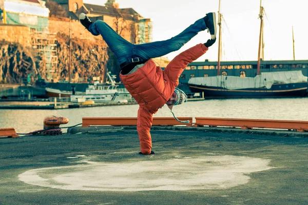 Young guy dancing break dance on the waterfront in an orange jacket in winter. Horizontally framed shot.