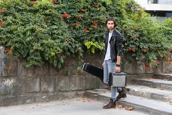 Street musician holding a case with a guitar and amplifier and looking into the distance. Vagrant lifestyle. Playing to make money a living. Unemployed musician. Future rock star. Horizontally framed shot.