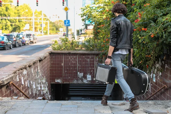 Street musician holding a case with a guitar and amplifier. Descends into the underground passage. Vagrant lifestyle. Playing to make money a living. Unemployed musician. Future rock star. Horizontally framed shot.