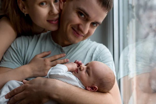 Mężczyznę trzymającego na rękach noworodka. Kobieta przytula człowieka od tyłu. Siedzi obok okna. Mama, Tata i dziecko. Portret młodej rodziny. Szczęśliwe życie rodzinne. Urodził się człowiek. — Zdjęcie stockowe