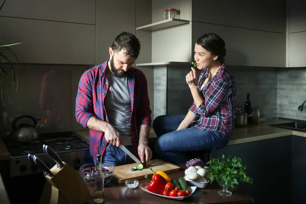 Beautiful young couple in kitchen at home while cooking healthy food. Man is cuts salad. Woman sits on table and eating parsley. Scene from family life. — Stock Photo, Image