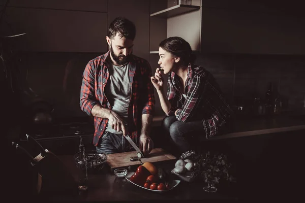 Beautiful young couple in kitchen at home while cooking healthy food. Man is cuts salad. Woman sits on table and eating a cucumber. Scene from family life. — Stock Photo, Image