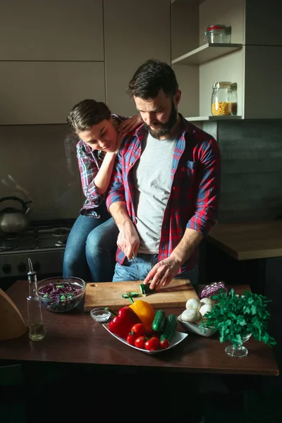 Beautiful young couple in kitchen at home while cooking healthy food. Man is cuts salad. Woman sits on table and hugs man from behind. Scene from family life. — Stock Photo, Image