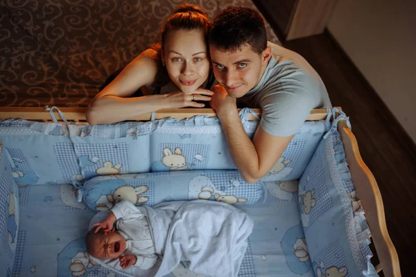 Man en een vrouw in de buurt van hun pasgeboren baby. Ouders kijken naar de camera. De kreten van de jongen in zijn wieg. Moeder, vader en baby. Portret van jonge gezin. Gezinsleven. De mens werd geboren. — Stockfoto