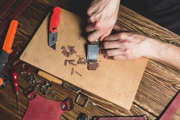 마스터 칼과 가죽 조각의 그의 손에 보유 하고있다. 도구 및 액세서리와 함께 흩어져 갈색 나무 테이블에. — 스톡 사진