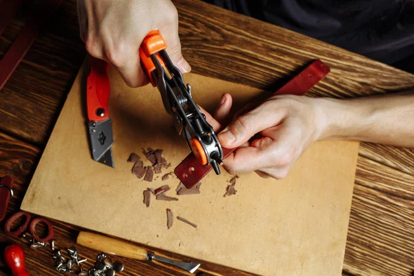 구멍 펀치와 가죽의 한 조각 들고 마스터. 도구 및 액세서리와 함께 흩어져 갈색 나무 테이블에. — 스톡 사진