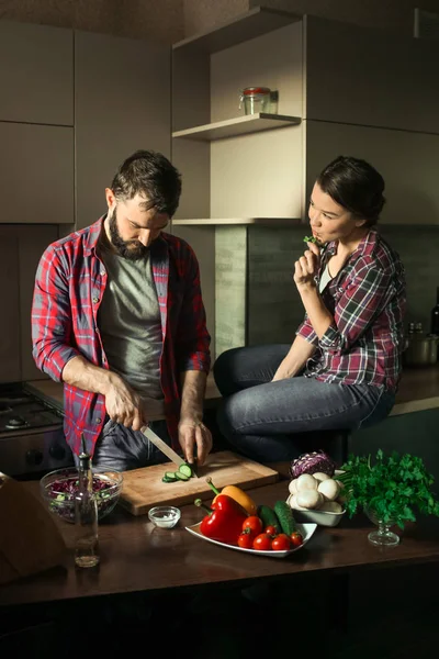 Belo casal jovem na cozinha em casa enquanto cozinha comida saudável. O homem corta salada. A mulher senta-se à mesa e come salsa. Cena da vida familiar . — Fotografia de Stock