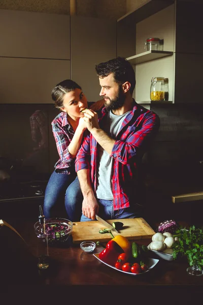 Beautiful young couple in kitchen at home while cooking healthy food. Husband cuts salad and feeds his wife cucumber. Woman looks at the frame. Scene from family life.