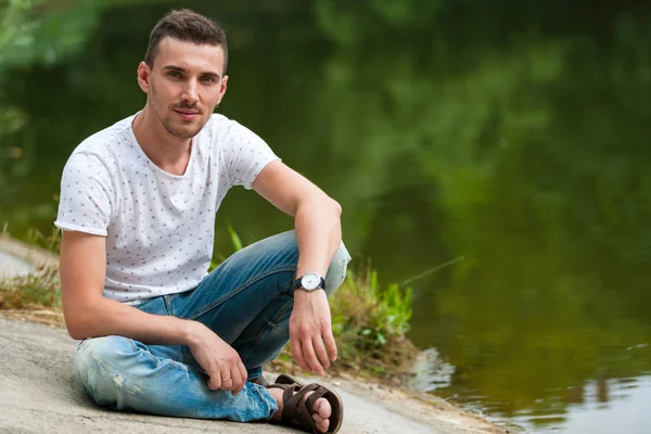 若いハンサムな男の湖の横に座っている。水は木を反射する. — ストック写真