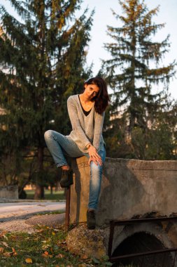 Seksi esmer bir meydan okuyan poz oturur. Arka planda yeşil Firs.