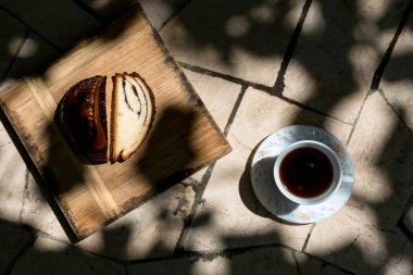 Bir fincan sade kahve ve bir rulo haşhaş tohumu. Doğal taşın zemininde. Yukarıdan görüntüle. Yapraklardan derin gölgeler.