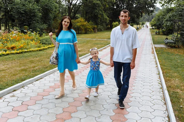 Щаслива сім'я йде по шляху тримаючи руки і проводячи час у прекрасному зеленому парку. Жінка вагітна . — стокове фото