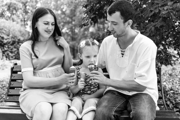 Батько, мати і дочка сидять на лавці і їдять морозиво. Чоловік ділиться морозивом з дівчиною. Жінка вагітна . — стокове фото