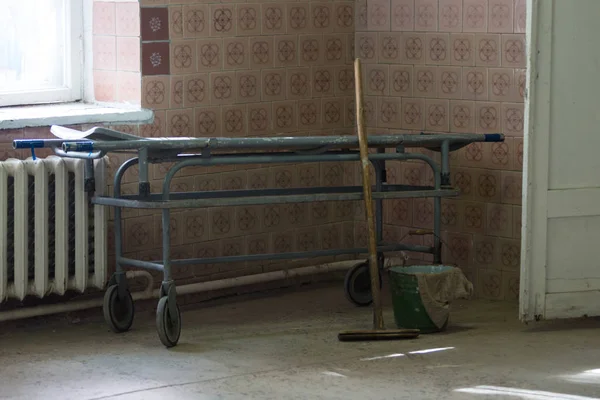 Cama de maca velha no corredor do hospital. Balde de madeira esfregão e pano. Um horrível hospital velho ou morgue . — Fotografia de Stock
