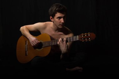Yakışıklı genç müzisyen gitar çalıyor. Çıplak gövde. Siyah arka planda.