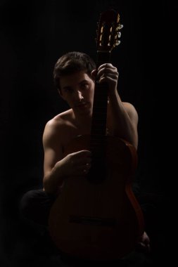 Yakışıklı bir genç müzisyen elinde bir gitar tutar. Çıplak gövde. Siyah arka planda.
