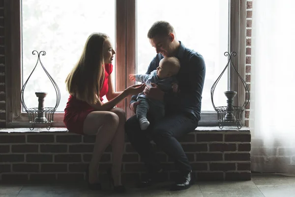 Rodzice i ich dziecko siedzą przy oknie. Mama, tata i dziecko. Portret młodej rodziny. Szczęśliwego życia rodzinnego. Człowiek się urodził.. — Zdjęcie stockowe