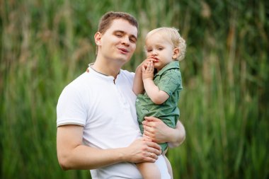 Baba küçük oğlunu kollarında tutuyor. Arka planda sazlıklar var. Parkta yürü. Yatay çerçeveli çekim.