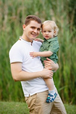 Baba küçük oğlunu kollarında tutuyor. Arka planda sazlıklar var. Parkta yürü. Dikey çerçevelenmiş çekim.