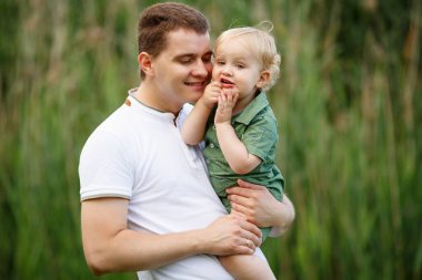 Baba küçük oğlunu kollarında tutuyor. Arka planda sazlıklar var. Parkta yürü. Yatay çerçeveli çekim.