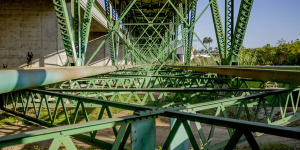 Support structure of a bridge in Oceanside CA. The structure under a bridge with the beams criss crossing in a geometric pattern. The sun shines brightly on the dirty and slightly rusty green metal.