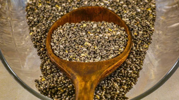 Chia seeds and wooden spoon inside a glass jar. Chia seeds and brown wooden spoon inside a glass jar lying on a shiny surface. Chia seeds are oval shaped and gray colored with black and white spots.