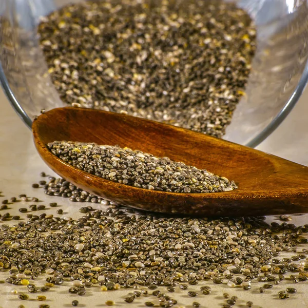 Chia seeds spilled from glass with wooden spoon. Oval and gray chia seeds spilled out from a glass container on to a shiny surface. Some of the chia seeds are on a brown wooden spoon.