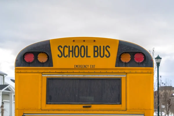Close up of the back side of a yellow school bus against homes and cloudy sky