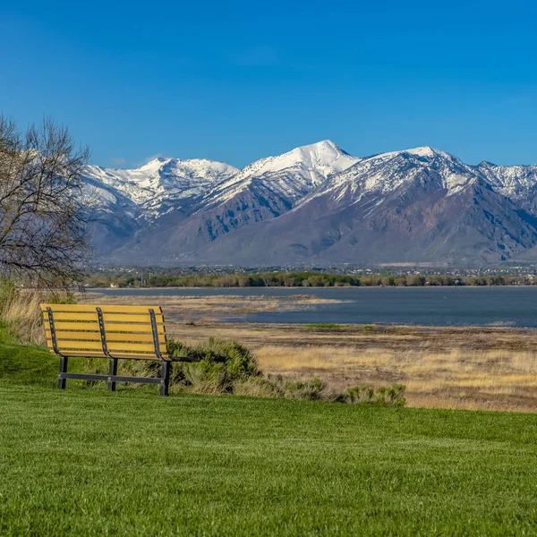 Čtvercová lavička na svěžím poli s výhledem na jezero a nad sněhem pokryté hory — Stock fotografie