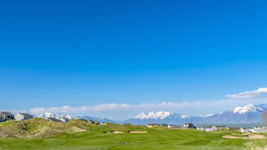 Panoramik geniş çimenli alan, eğimli döşeli bir yol ve yamaçların ötesinde evler
