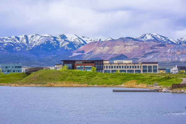 Buildings and snowy mountain beyond the grassy shore of a lake on a cloudy day