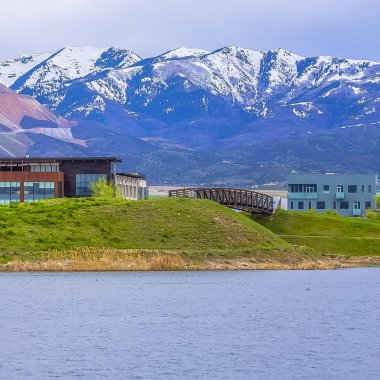 Kare çerçeve binalar ve bulutlu bir gün bir gölün çimenli kıyı ötesinde karlı dağ